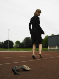 Sophie running on race track in black suit, her broken shoe in foreground