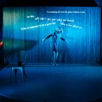sophie cringes on a blue lit stage as if scared by an unseen enemy. behind her text is projected ,phrases such as 'why cna't you just leave me alone' and 'i'm running all over the place without music'. behind sophie is a slatted curtain. she stands on a lip shaped mat on the stage. there is a simple metal chair and sci fi style lights at the side of the stage. sophie wears a dress silver leggings and has bobbed hair that flies out as she swivels.