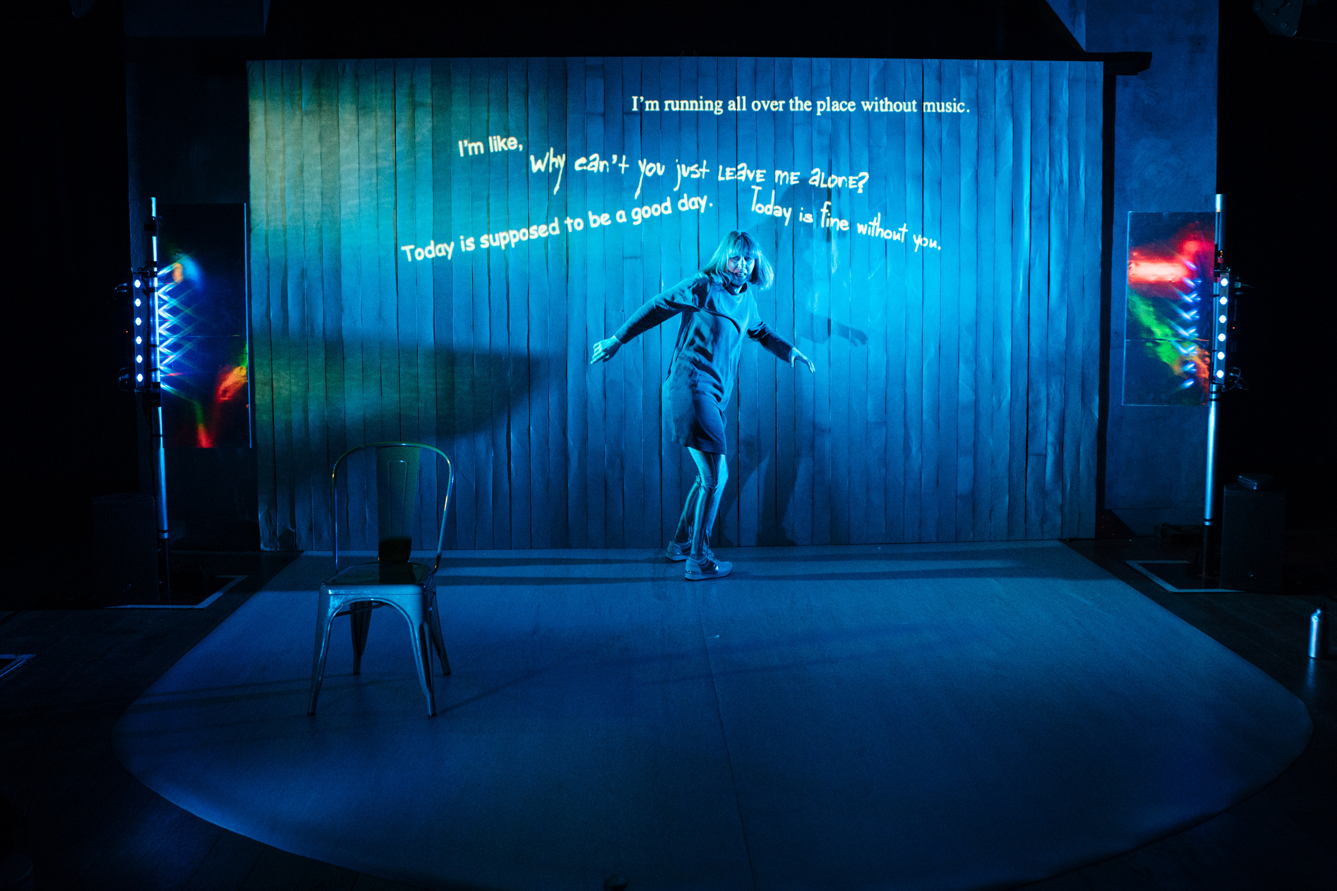 sophie cringes on a blue lit stage as if scared by an unseen enemy. behind her text is projected ,phrases such as 'why cna't you just leave me alone' and 'i'm running all over the place without music'. behind sophie is a slatted curtain. she stands on a lip shaped mat on the stage. there is a simple metal chair and sci fi style lights at the side of the stage. sophie wears a dress silver leggings and has bobbed hair that flies out as she swivels.