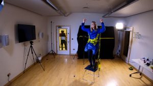 Sophie a white woman dressed in blue and also bin bags and warning yellow distance tape stands on a chair in a studio room roaring in triumph, being filmed through a glass door by a man. 